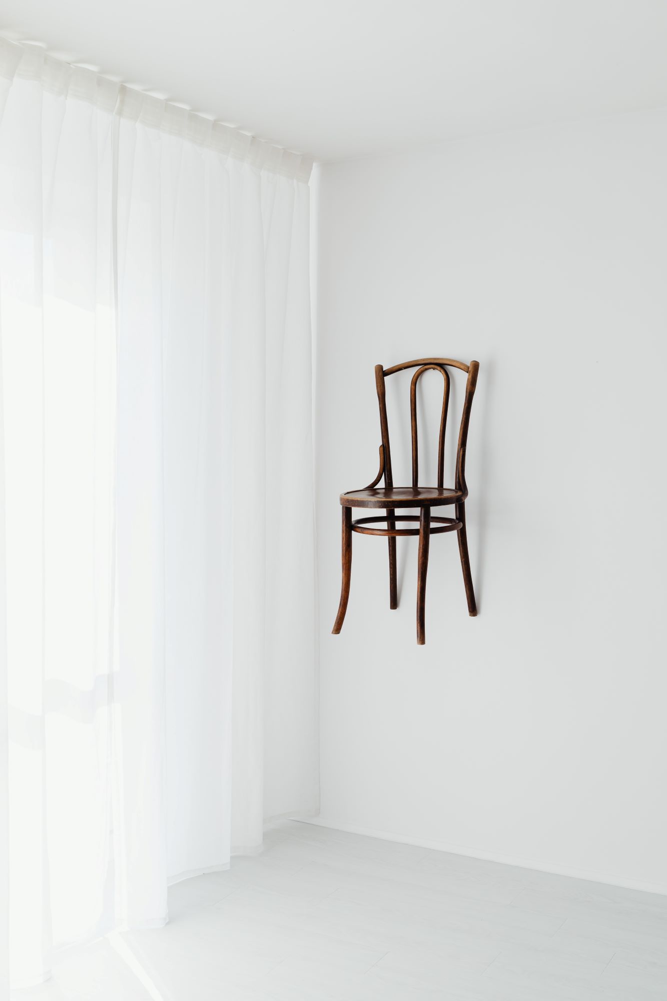 An einer weißen Wand mit einer weißen Gardine hängt an einer brauner Holzstuhl