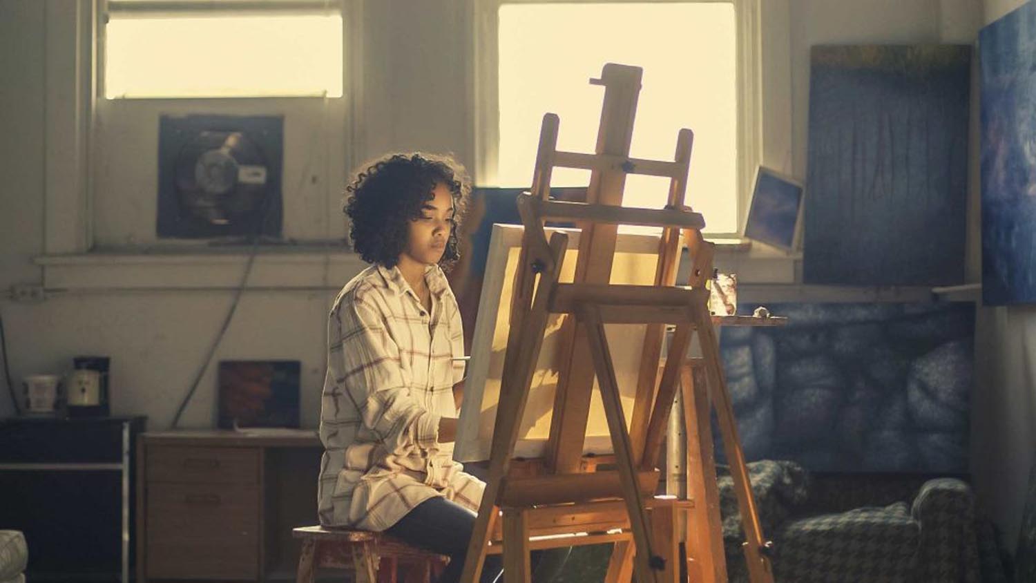 Eine schwarze Frau sitzt in einem Atelier vor einer Staffelei und malt