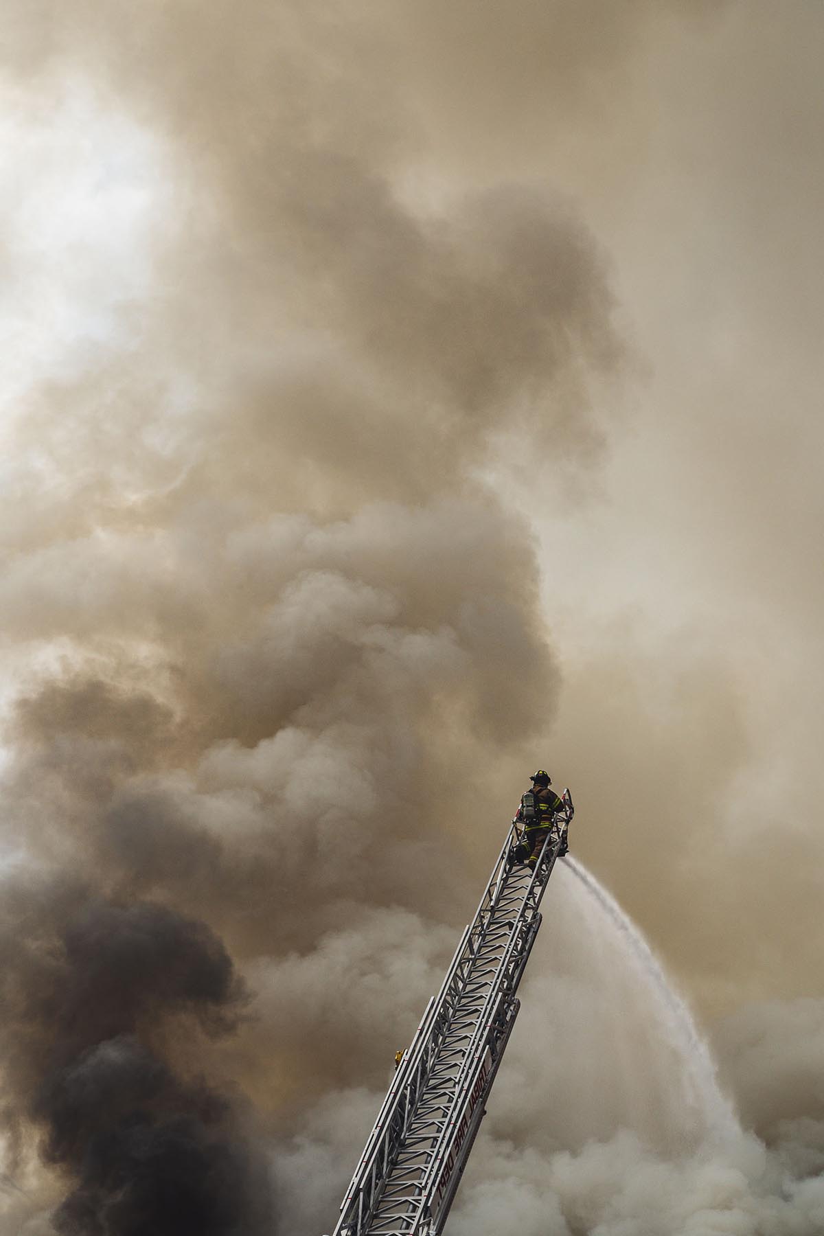 Foto eines stark verrauchten Himmels mit einem Feuerwerhmann auf einer Hochleiter, der verscuht zu löschen