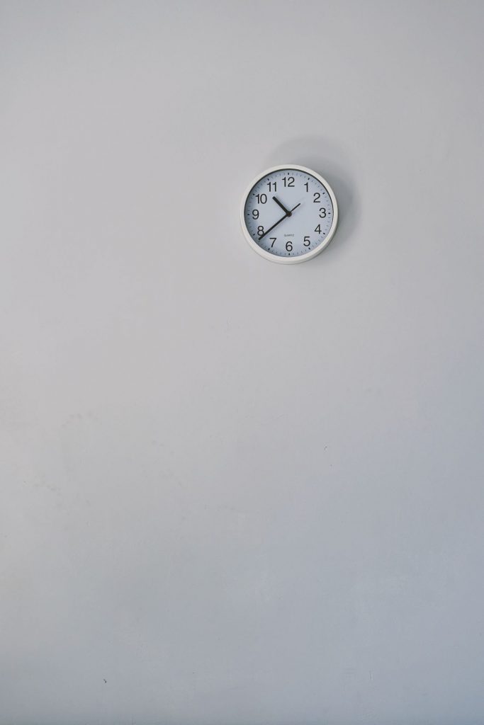 An einer langen weißen Wand hängt eine weiße Uhr die zwanzig vor elf Uhr anzeigt