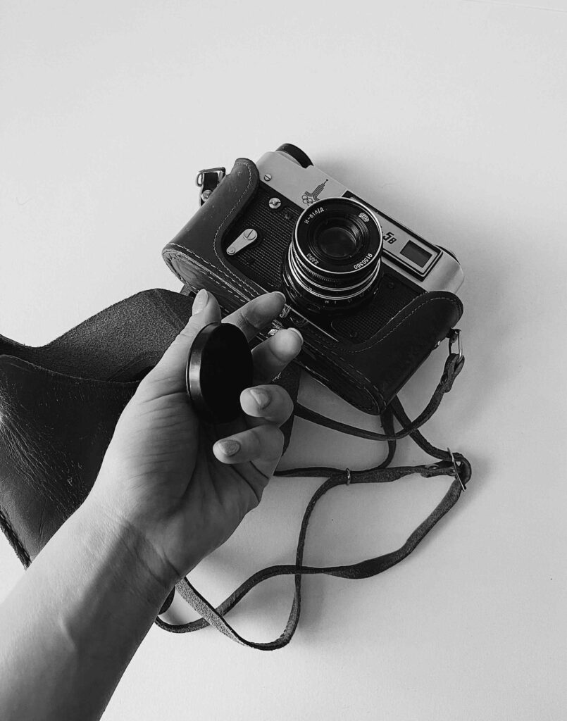 Schwarz-Weiß-Foto einer analogen Kamera und einer Hand, die den Deckel vom Objektiv nimmt