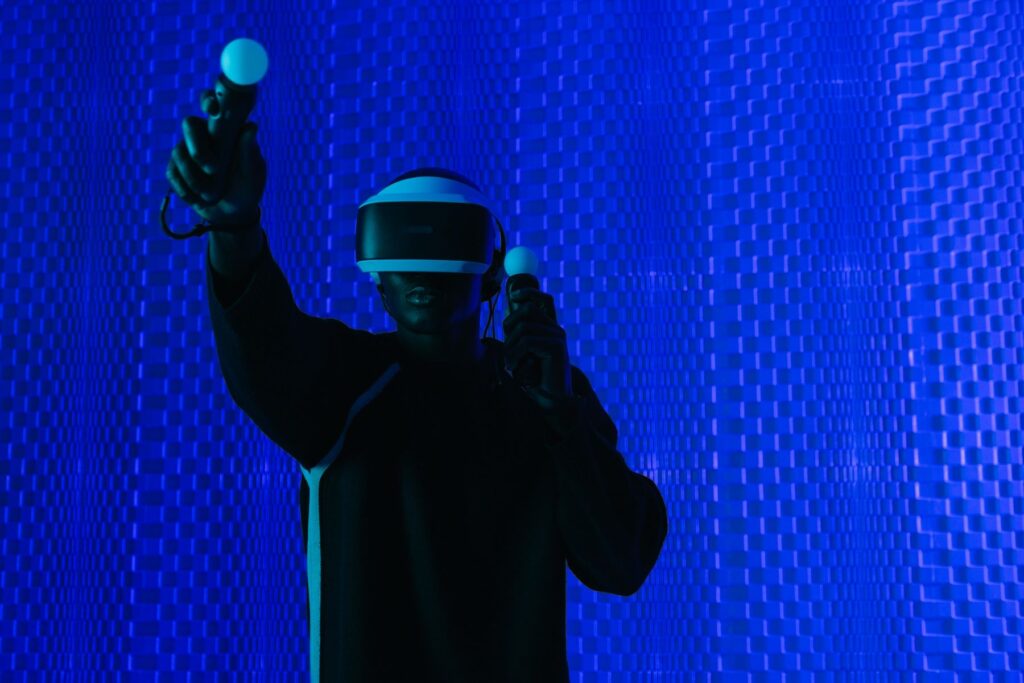 Ein schwarzer Mann steht mit Virtual Reality Brille und Joysticks vor einener leuchtenden blauen Wand