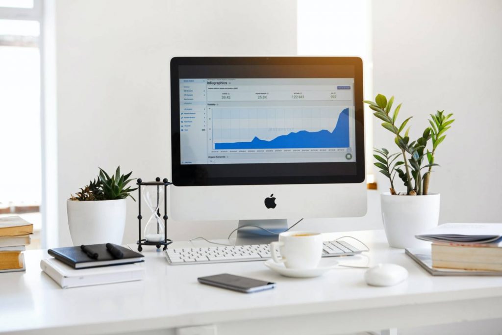Ein großer iMac mit einer Infografik auf dem Display steht auf einem weißen Schreibtisch mit Grünpflanzen Kaffeetasse iPhone Büchern und Notizbüchern sowie einer Sanduhr