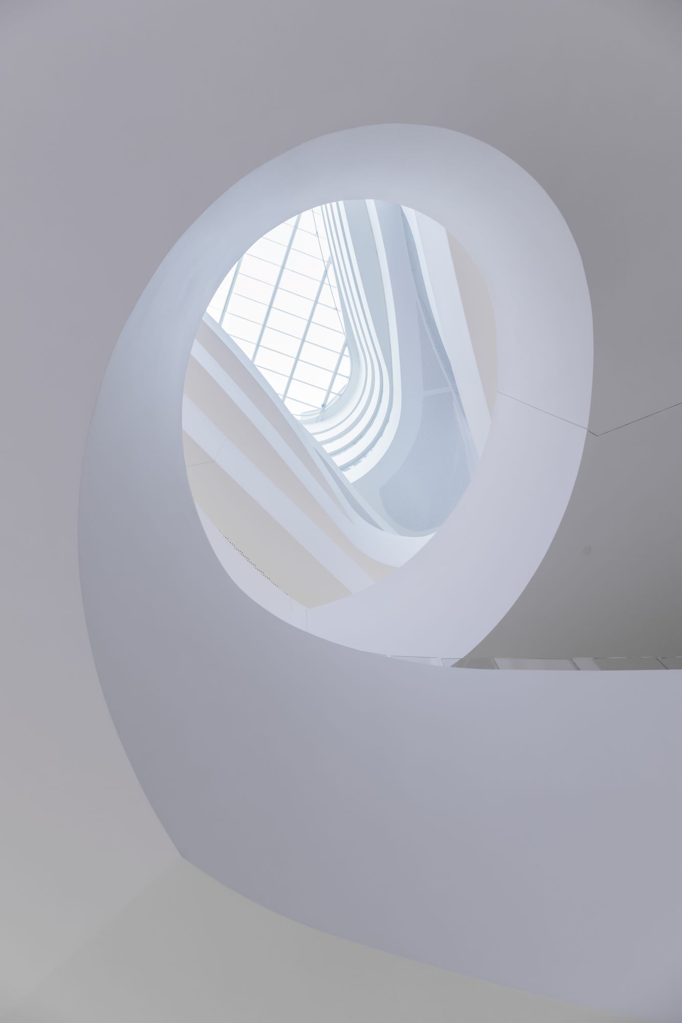 Von unten nach oben fotografiertes weißes rund zulaufendes Treppenhaus mit zeitloser Architektur und Glasdach