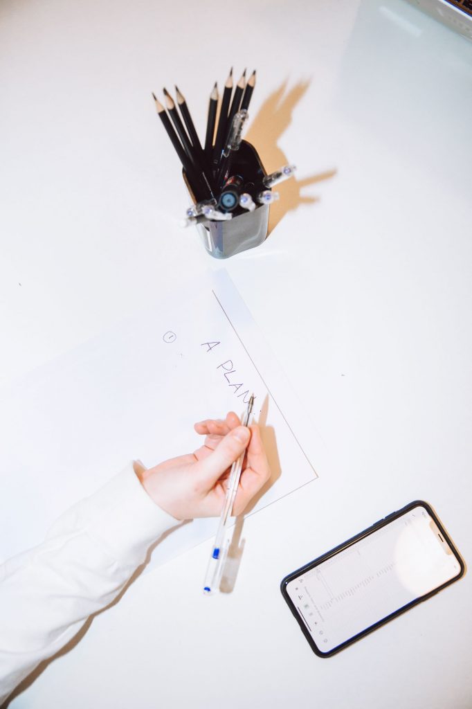 Auf einem weißen Tisch steht ein Gefäß mit schwarzen Stiften eine Frauenhand schreibt Pläne auf einen Zettel neben ihr lieht ein schwarzes iPhone