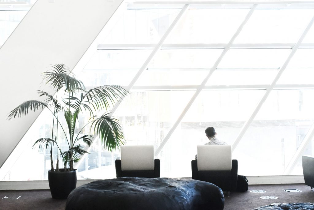 Ein helles Dachgeschoss in einem Bürogebäude zeigt einen Mann der auf einem Sessel sitzt an dem seine Laptoptasche lehnt davor Sitzinseln und eine Palme