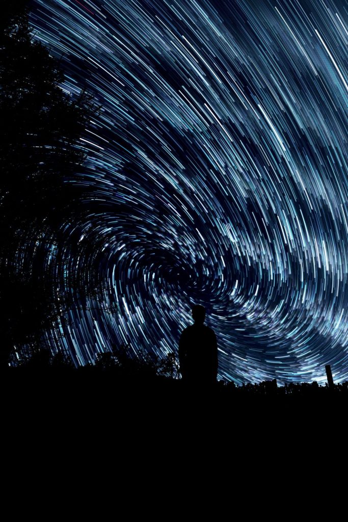 In tiefer Nacht steht ein Mensch auf einer Weide mit Baum und blickt auf verschwommene Sterne und Lichtkreise