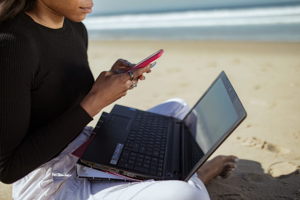 Eine Frau sitzt am Strand und hat ein Notebook auf ihrem Schoß und hält ein Handy in ihrer Hand