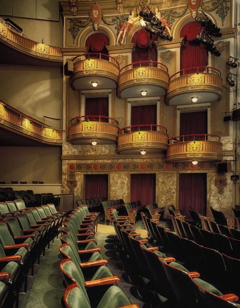 Ausschnitt eines Fotos von einem Theatersaal mit Samtklappsitzen, Balkonen und Hochrängen und aufwendiger Wandverzierung