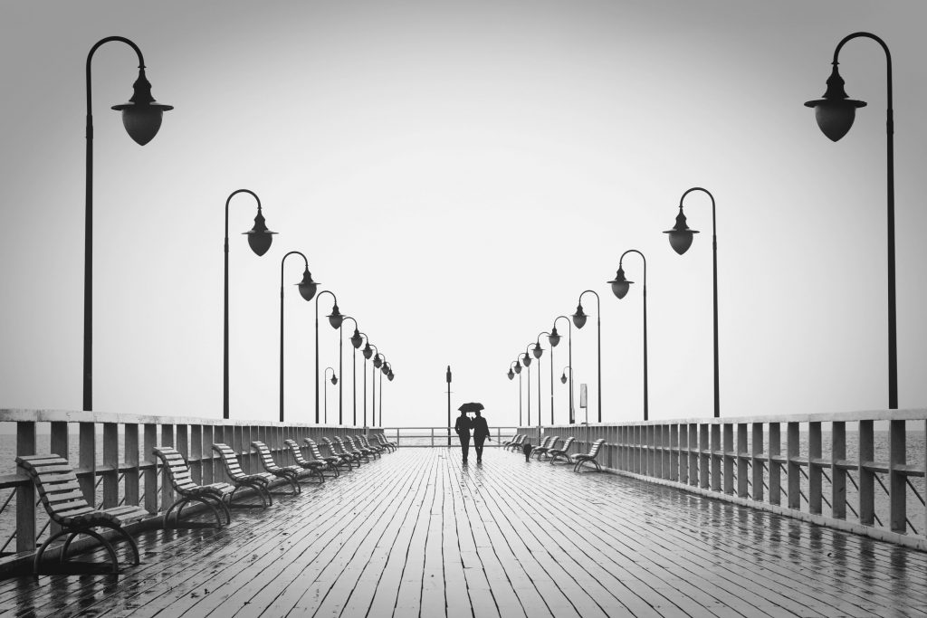 Schwarzweiß Foto von einem Paar das mit Hund und Regenschirm eine verlassene Promenade zum Meer entlang schlendert