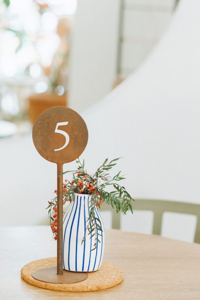 Auf einem runden Holztisch steht eine blauweiß gestreifte Vase mit Pistazienzweigen und Beeren zusammen mit dem Nummernschild 5 und davor ein grüner Stuhl