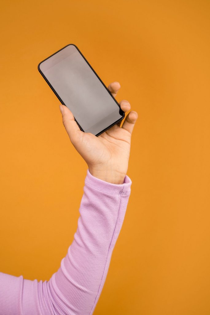vor einem knallig orangenen Hintergrund ist der Arm einer Frau in einem fliederfarbenen Pulli zu sehen mit einem Mobiltelefon in der Hand