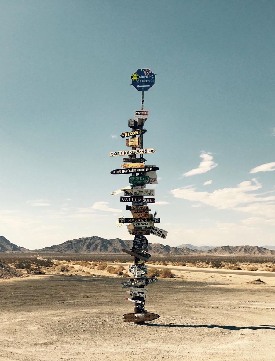 Mitten in der Wüste steht ein Metallpfahl mit unzähligen Schildern die in alle Richtungen zeigen zu Orten und Sehenswürdigkeiten