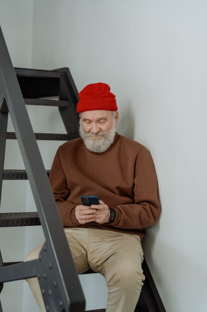 Ein älterer Mann mit roter Mütze sitzt auf einer Treppe und bedient sein Handy