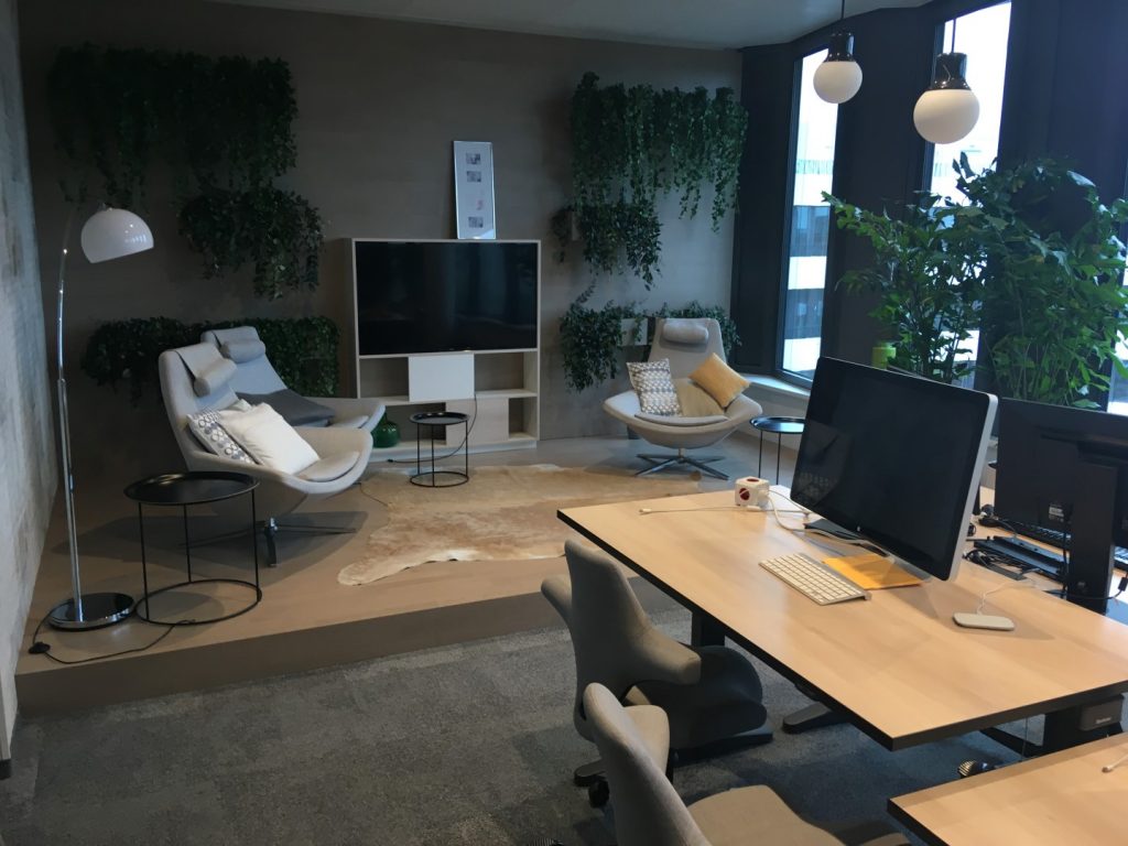Blick in eine gemischte Arbeitsplatzumgebung mit gemütlichen Sesseln Teppich Grünpflanzen verschiedenen Leuchten und klassischen höhenverstellbaren Schreibtischen mit großen Monitoren