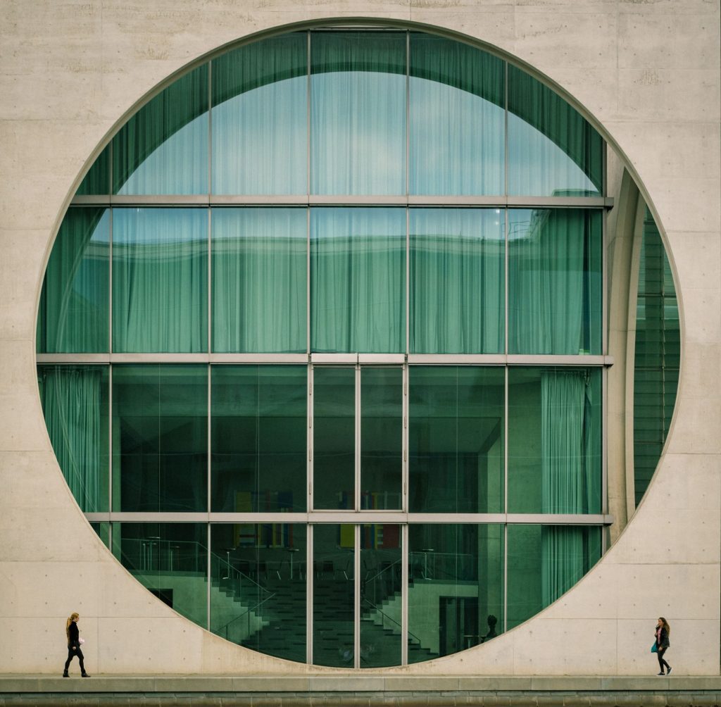 Eine beige Betonhausfassade mit einem großen Kreissauschnitt gibt den Blick frei auf ein Gebäude mit zahlreichen Fenstern zwei Frauen laufen sich davor entgegen