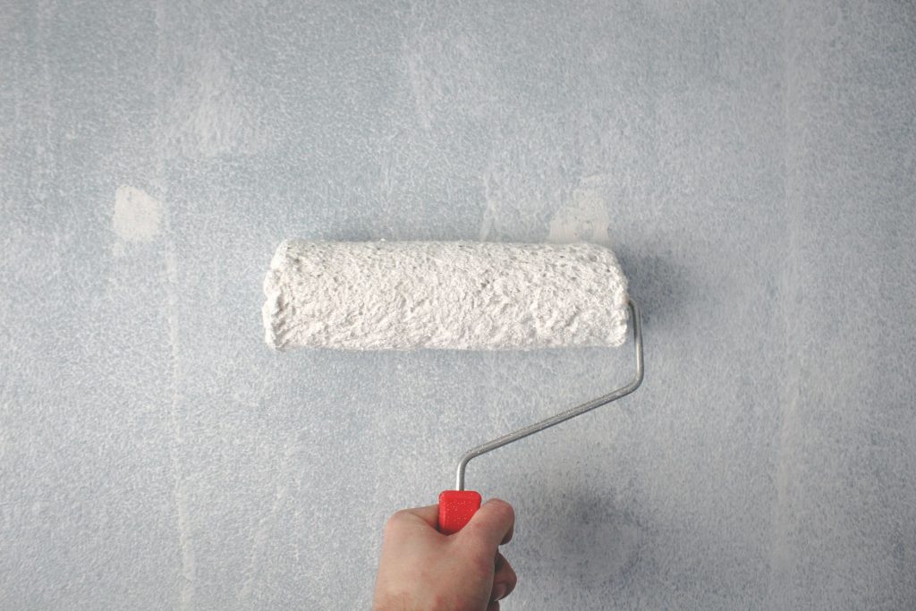 Eine Hand hält eine dicke Farbrolle mit weißer Wandfarbe und streicht eine Wand