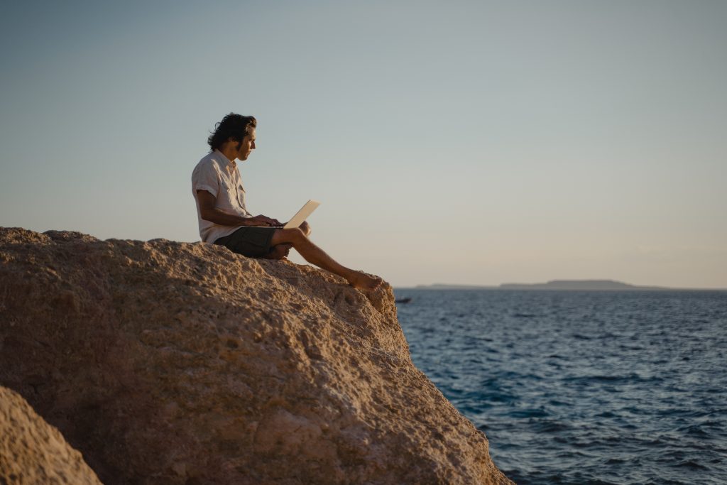 Weißer Mann sitzt auf einem Felsen am Meer und hat sein Notebook auf dem Schoß