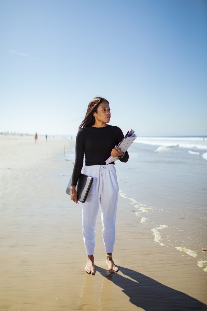 Eine schwarze Frau in Freizeitkleidung steht barfuß mit Aktentasche und Unterlagen am Strand im Sand