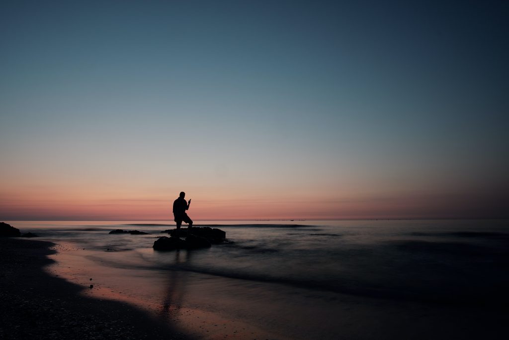 Bei Sonnenuntergang sieht man in der Ferne einen Mann auf einem Stein am Strand im Meer stehen