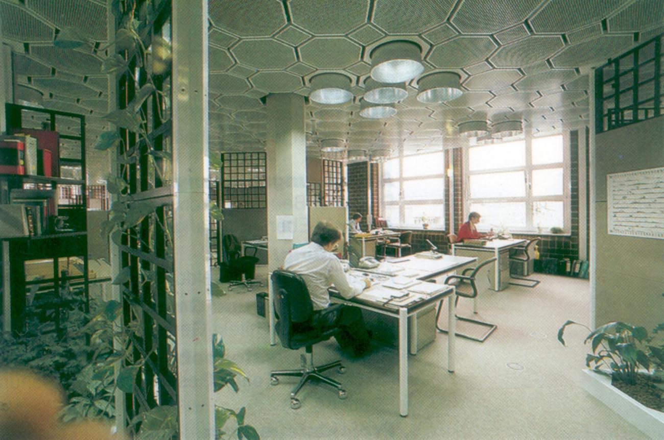 Auf einem Foto aus den Achtziger Jahren sitzen zwei Männer und eine Frau an ihren Schreibtischen auf einer großen geteilten Fläche die von Gitterwänden mit Grünpflanzen getrennt sind