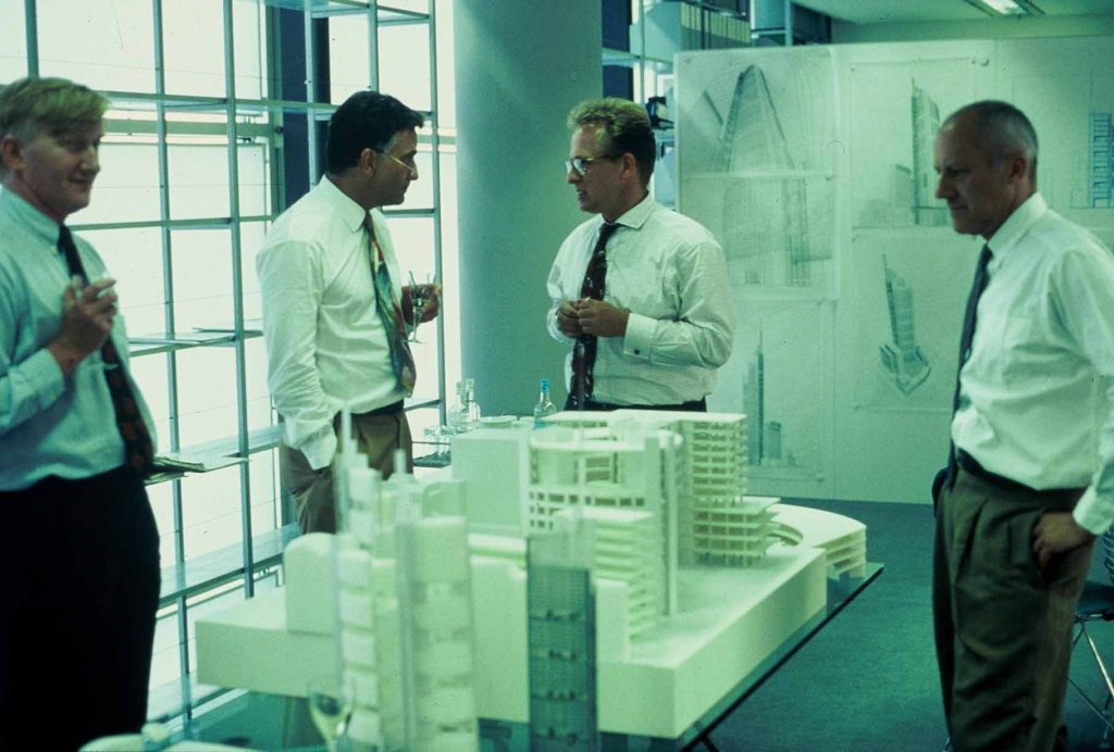 Auf einem Foto aus den Siebzigern stehen vier Männer mit Sektgläsern um einen Modellbau eines großen Geschäftsgebäudes hinter ihnen befinden sich Skizzen an der Wand