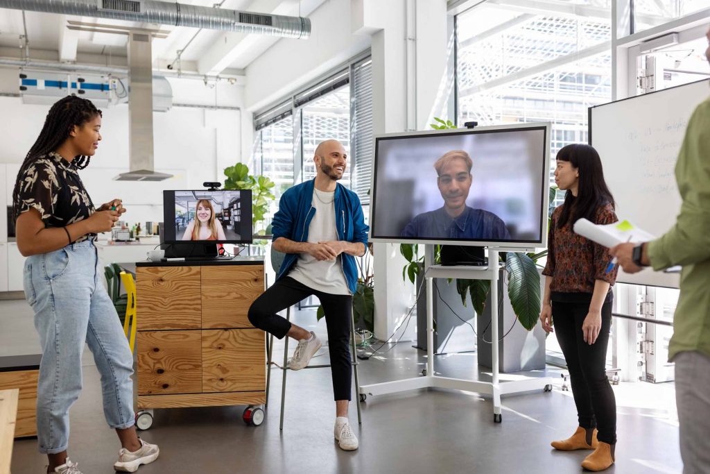 Auf einer industriellen Bürofläche die von Baustellen umgeben ist stehen zwei Männer zwei Frauen ein kleiner Bildschirm und ein großer Flatscreen über die Teilnehmer digital zugeschaltet sind