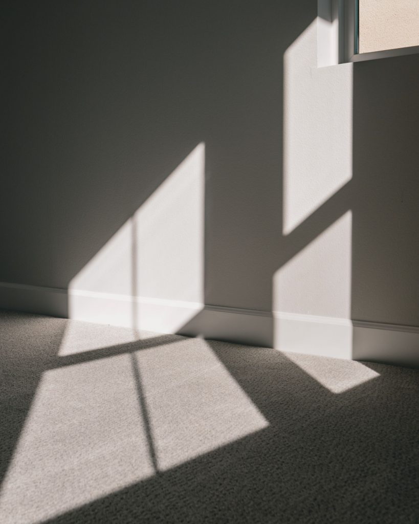 Ausschnitt eines Raumes, das einen grauen Teppich und eine hellgraue Wand mit Fenster-Ansatz zeigt, durch das Sonnenlicht einfällt und für ein Lichtspiel sorgt