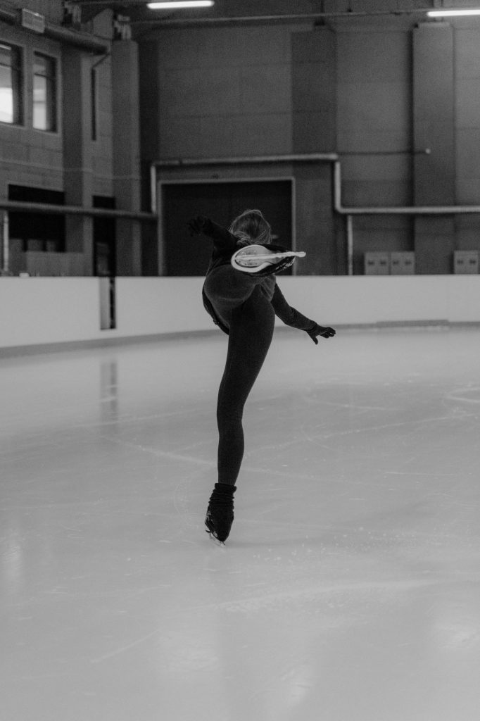 Schwarzweiß Foto einer schwarz gekleideten Eiskunstläuferin auf der glänzen Eisfläche in einem leeren Raum