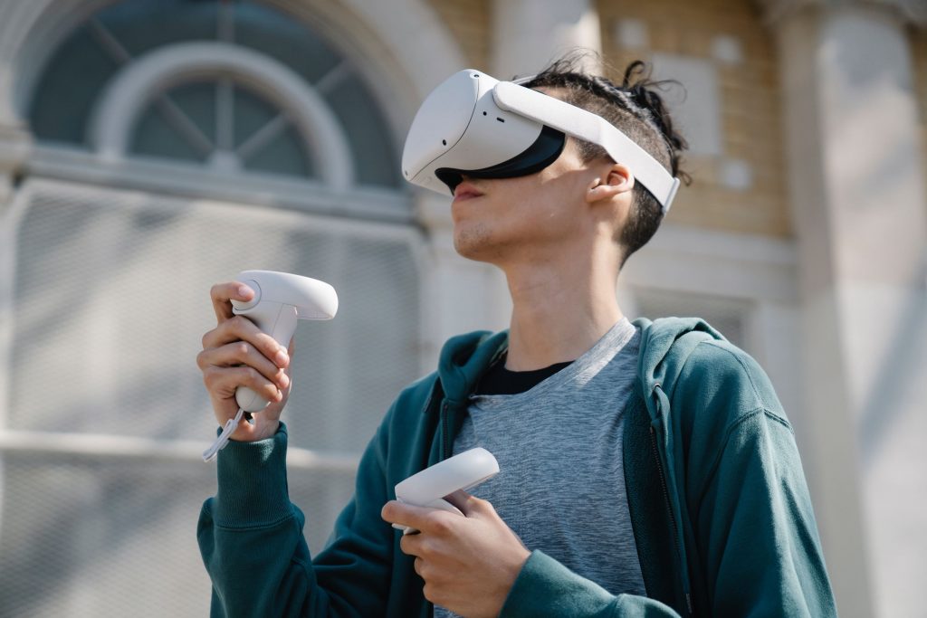 Ein junger Mann steht vor einem barocken Gebäude und trägt eine Virtual Reality Brille während er in jeder Hand je einen Controller hält