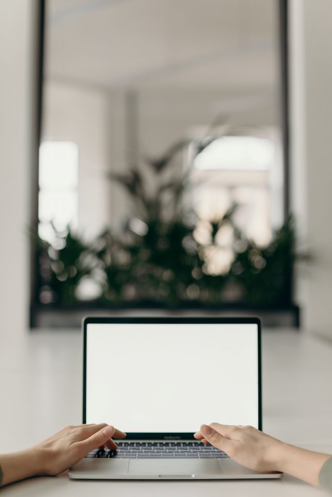 In einem weißen Raum mit Fenster in einen weiteren Raum stehen eine Grünpflanze und MacBook wird von zwei Frauenhänden benutzt