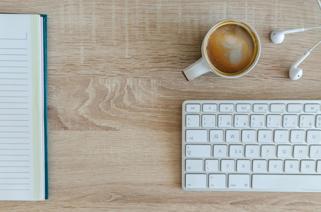 Auf einem Holztisch stehen eine Tasse Kaffee liegt ein Notizbuch eine Apple Tastatur und ein iPhone Kopfhörer