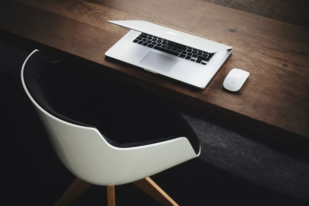 Weisser Schalenschreibtischstuhl mit Holzbeinen vor dunklem Holztisch mit MacBook und Mouse