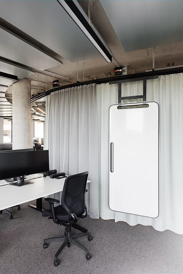 Ein offenes Büro mit einem höhenverstellbaren Tisch und einem Monitor und Stuhl. An einer Schiene vor einem Vorhang befindet sich ein verschiebbares Whiteboard.