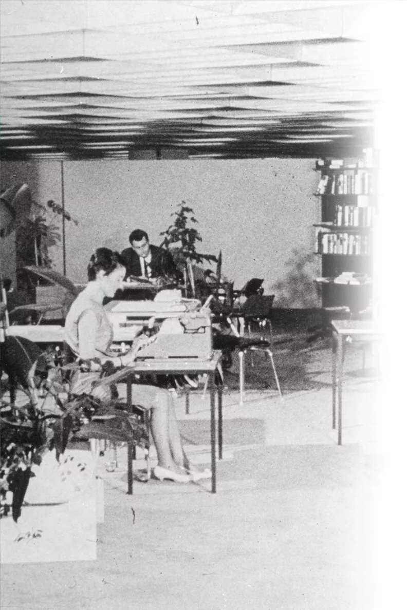 Größerer Ausschnitt eines unscharfen Schwarzweiß Fotos von einer alten Bürosituation mit einer Frau an der Schreibmaschine und zwei Männern die jeweils an ihren Schreibtischen sitzen