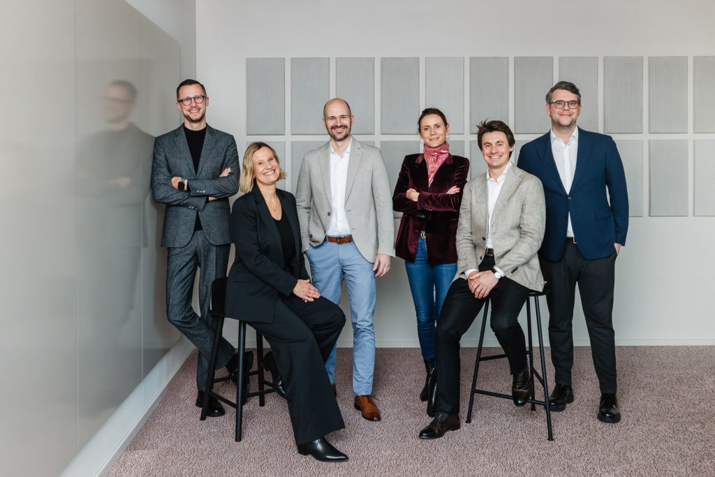 Eine Gruppe von Menschen steht vor einer Wand. Sie sind Business Casual gekleidet.