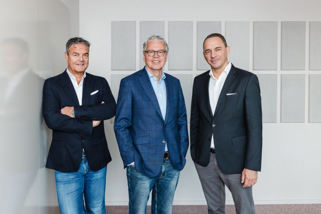 Drei Männer stehen vor einer Wand und blicken freundlich, aber entschlossen in die Kamera. Sie sind Business Casual gekleidet.