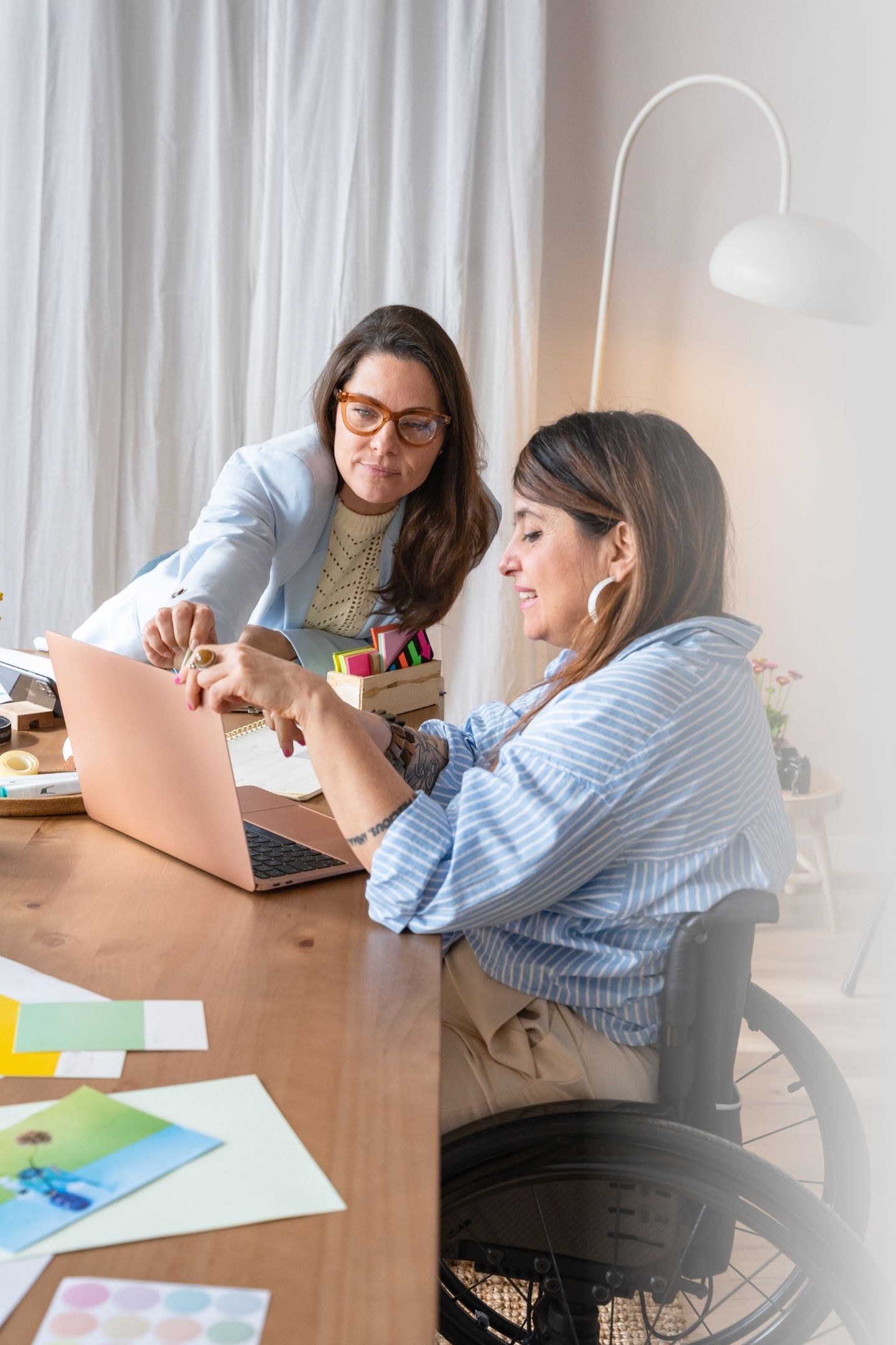 Zwei Frauen sitzen an einem Tisch und besprechen Arbeitsinhalte. Eine sitzt im Rollstuhl